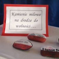Na stole ustawione są znicz i kamienie pomalowane w ważne historycznie daty oraz kartka z tytułem przedstawienia: 